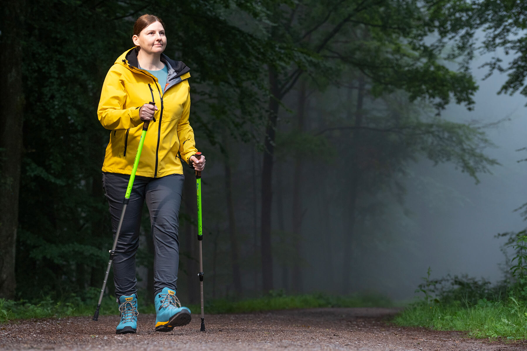 A woman in a yellow jacket hiking with poles through a misty forest trail