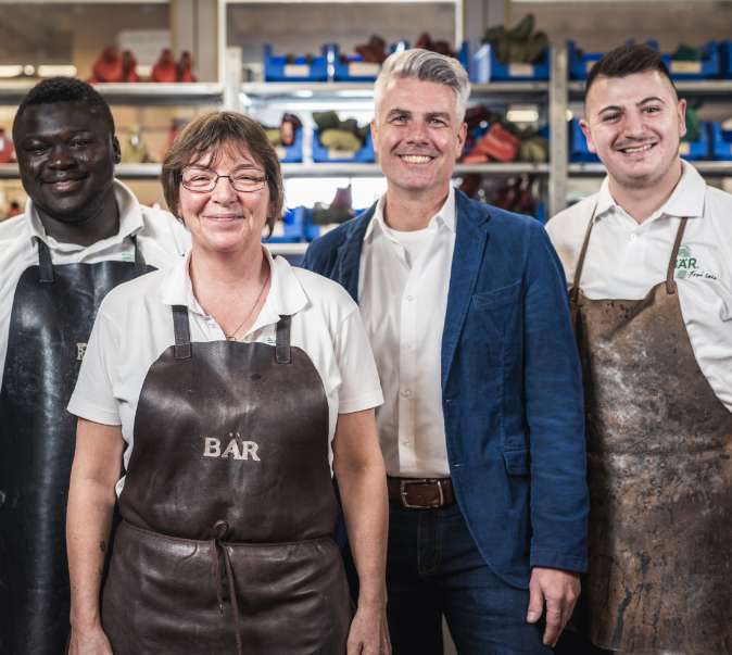 A group of four shoemakers, including three men and one woman, posed in a workshop setting, wearing aprons and smiling.