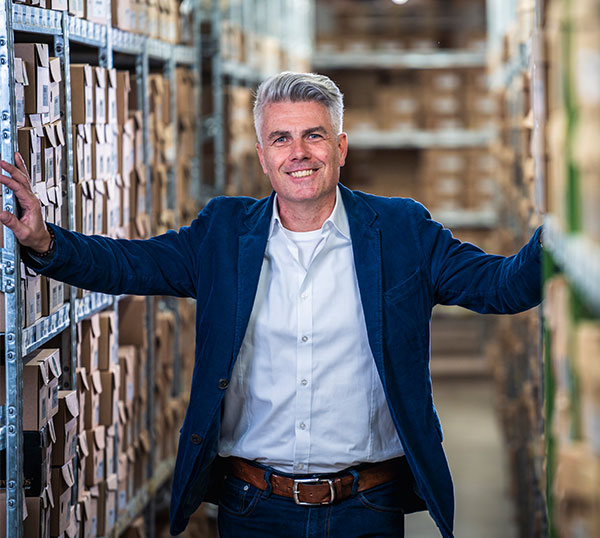 A smiling businessman with gray hair in a blue jacket stands confidently in a warehouse aisle surrounded by shelves of boxes.