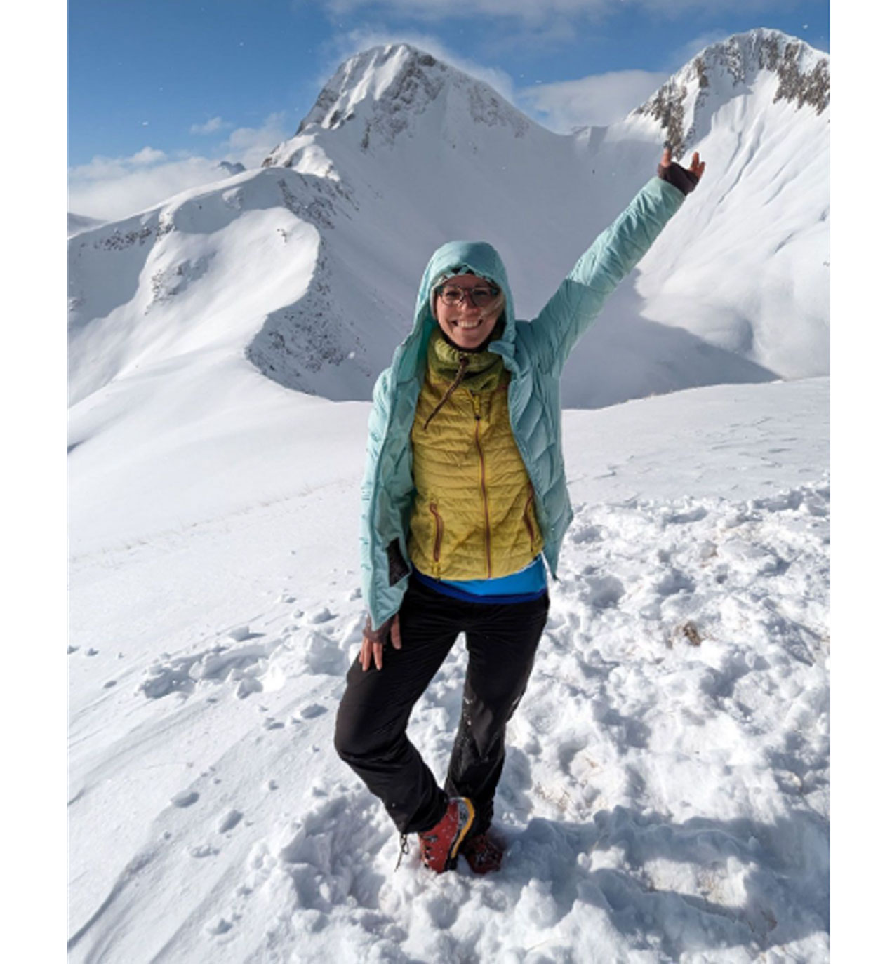 Woman in winter gear joyfully raises her arm while standing on a snowy mountain peak, clear blue sky in the background.