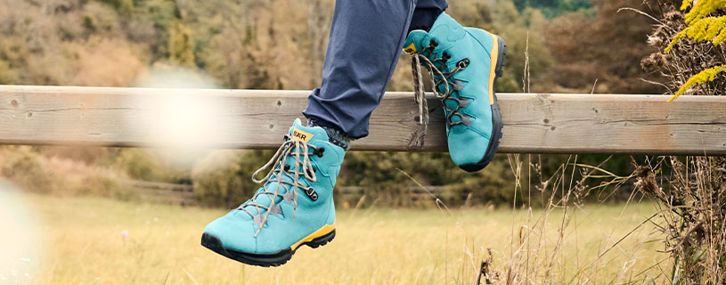 Person wearing blue hiking boots while sitting on a wooden fence in a grassy outdoor setting