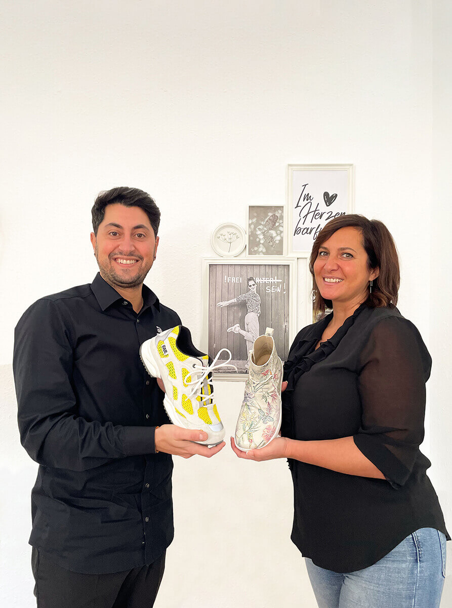 A man and a woman smiling and holding sneakers, standing in a room with artwork on the wall.