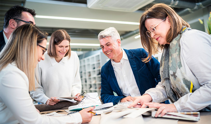 A group of five professionals discussing a design project with swatches and sketches in a bright and modern office space.