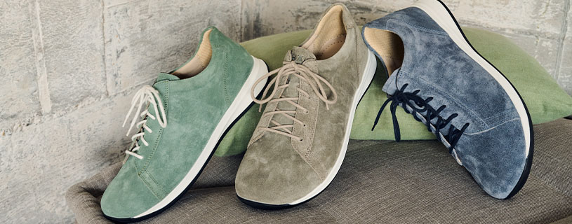 Three pairs of suede sneakers in green, beige, and blue displayed on a cushion against a rustic wall.