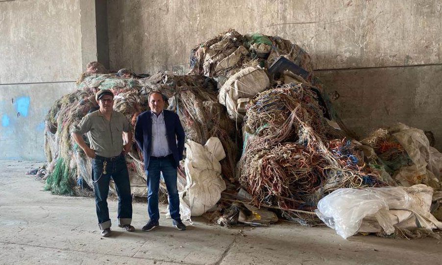 Two men stand beside a large pile of discarded fishing nets and ropes inside a warehouse environment