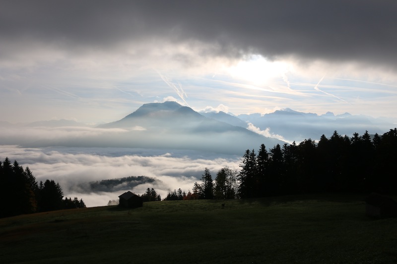 A serene mountain landscape with foggy valleys and a sun rising behind the clouds, surrounded by dark silhouettes of trees.