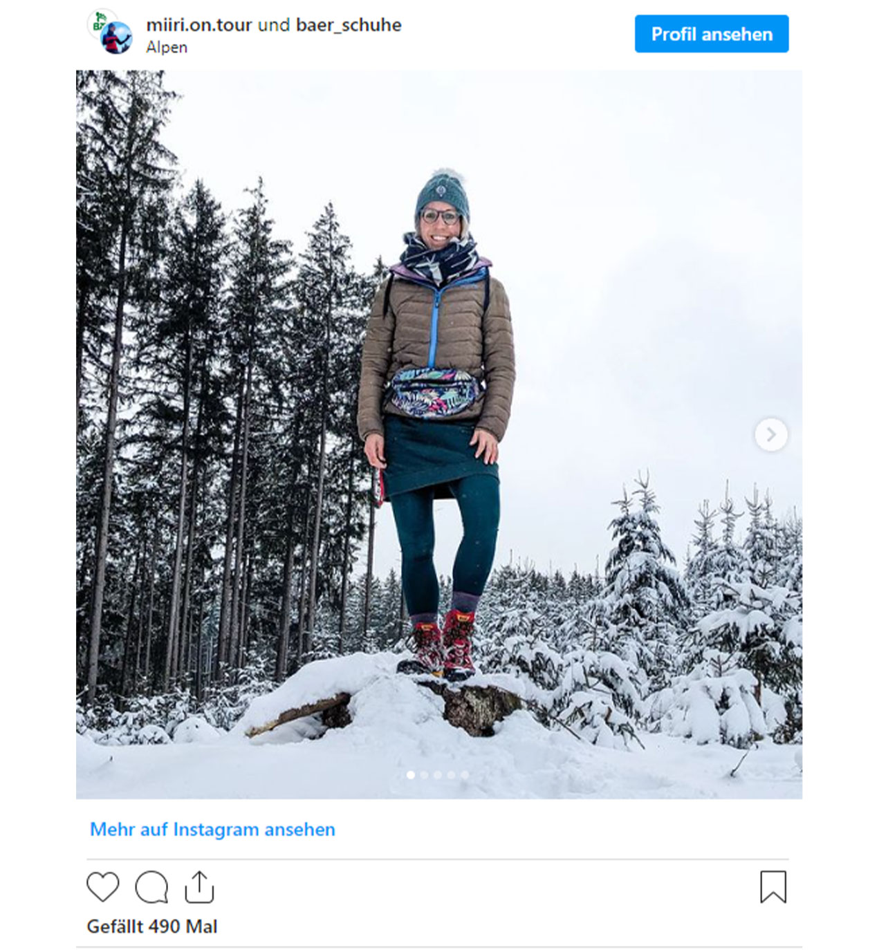 A woman wearing winter clothing stands atop a snow-covered stump, surrounded by tall trees in a snowy landscape.