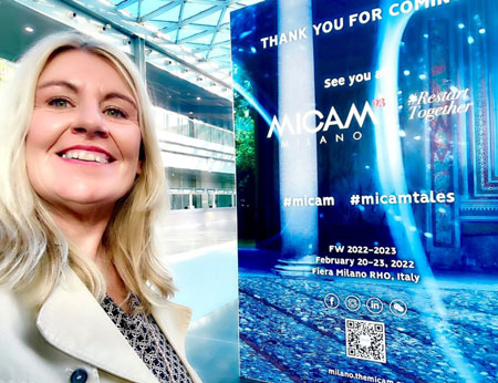 A woman smiling next to a MICAM Milano event poster.