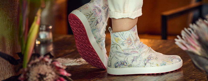 Person wearing floral patterned high-top shoes with white and pink colors, standing on a wooden floor with a flower nearby.