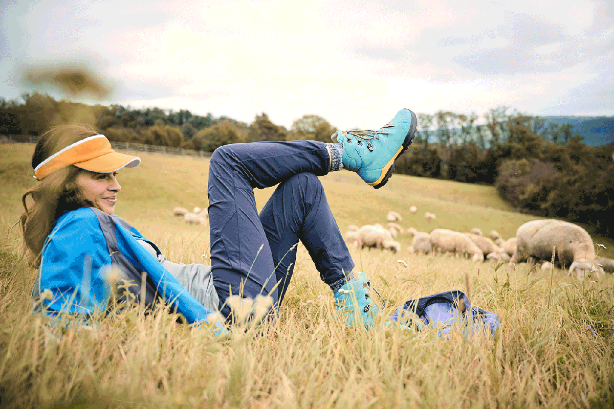 A woman in outdoor clothing and blue hiking boots relaxing in a field with grazing sheep.