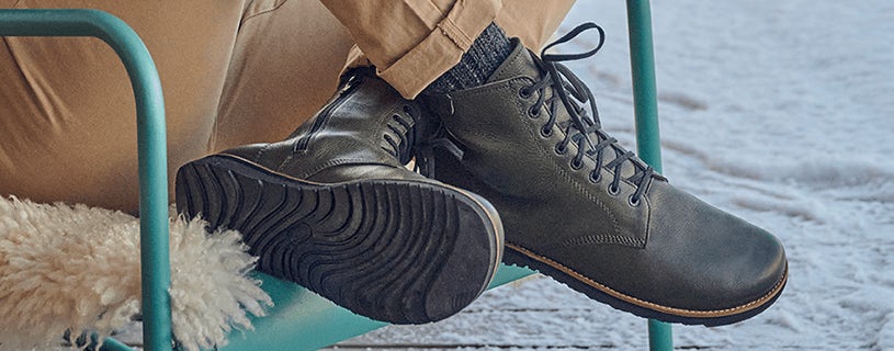 Person wearing cozy black leather boots with laced design and non-slip soles, sitting on a turquoise chair with a fuzzy cushion