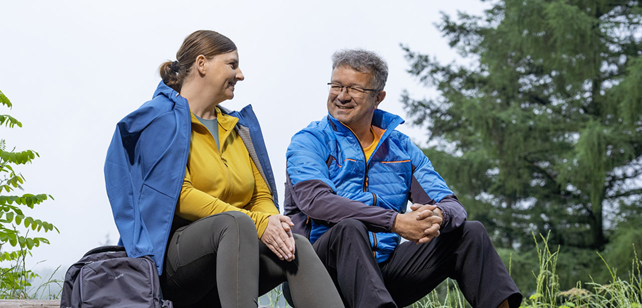 A man and a woman sitting on a log in a forest, wearing blue and yellow outdoor jackets, smiling at each other.