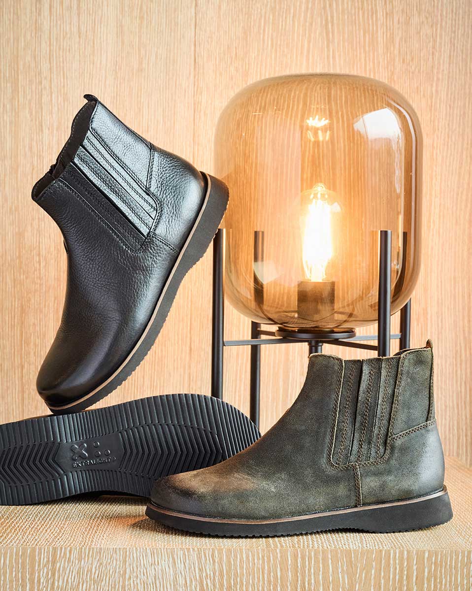 Two pairs of leather Chelsea boots displayed beside a modern lamp on a wooden surface.