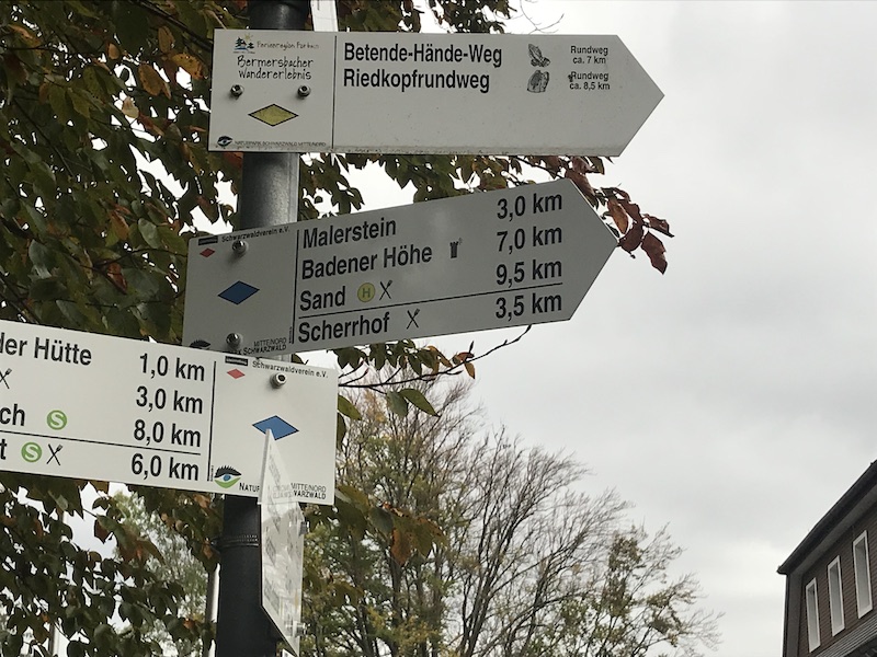 Direction signs for hiking trails, including Betende-Hände-Weg and Riedkopfrundweg, with various distances shown in a forested setting