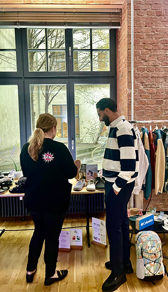 Two people browsing clothing and accessories in a boutique store with large windows and a brick wall backdrop.