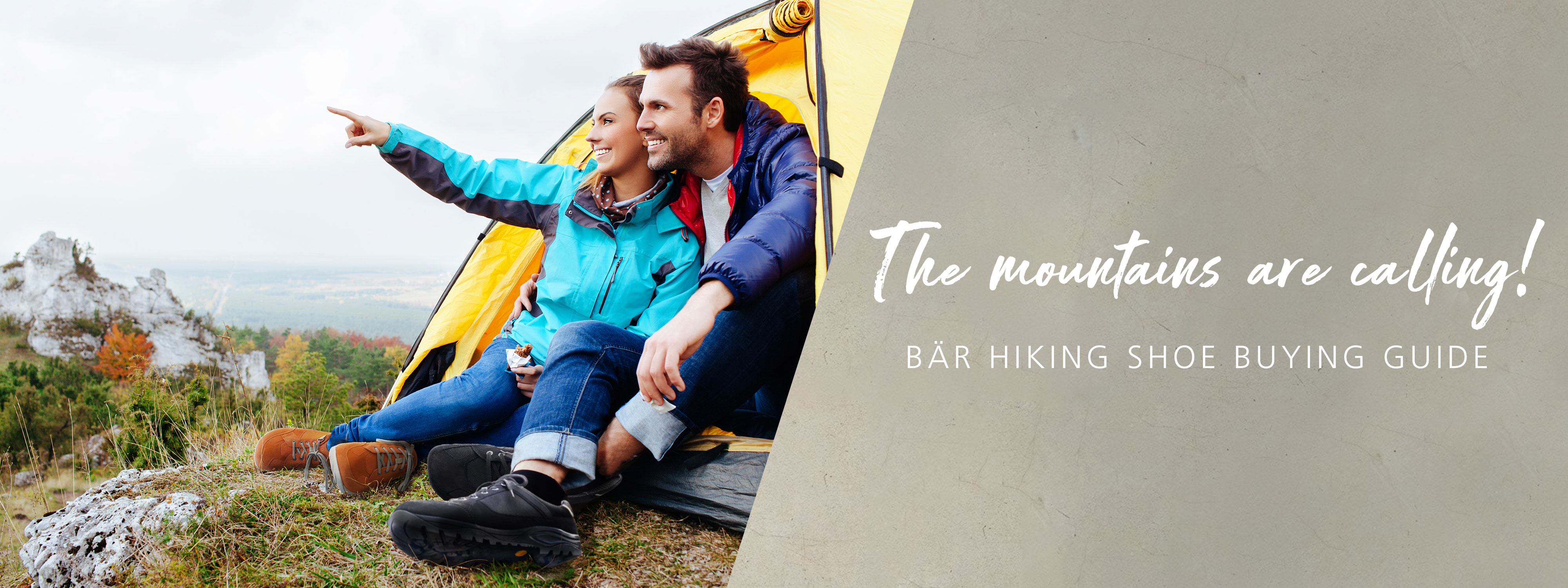 A happy couple sitting outside a yellow tent, pointing towards the mountains, with trees and rocky landscape in the background.