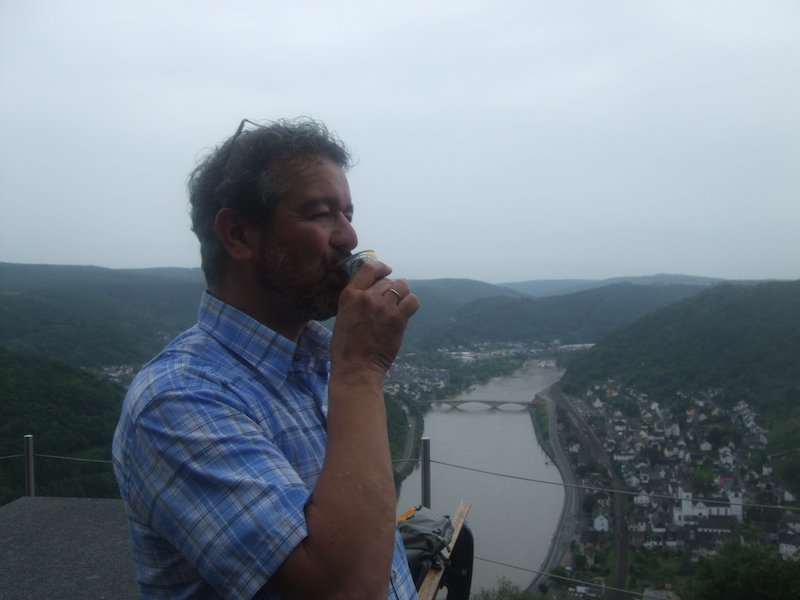 A man in a plaid shirt drinks from a can, overlooking a river and hilly landscape.