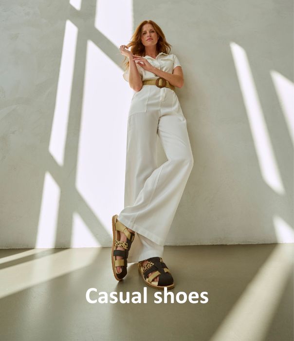 Woman leaning against wall in white outfit with casual sandals in sunlight