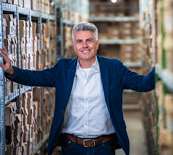 A man in a blue blazer smiling, standing in a warehouse aisle with shelves full of boxes